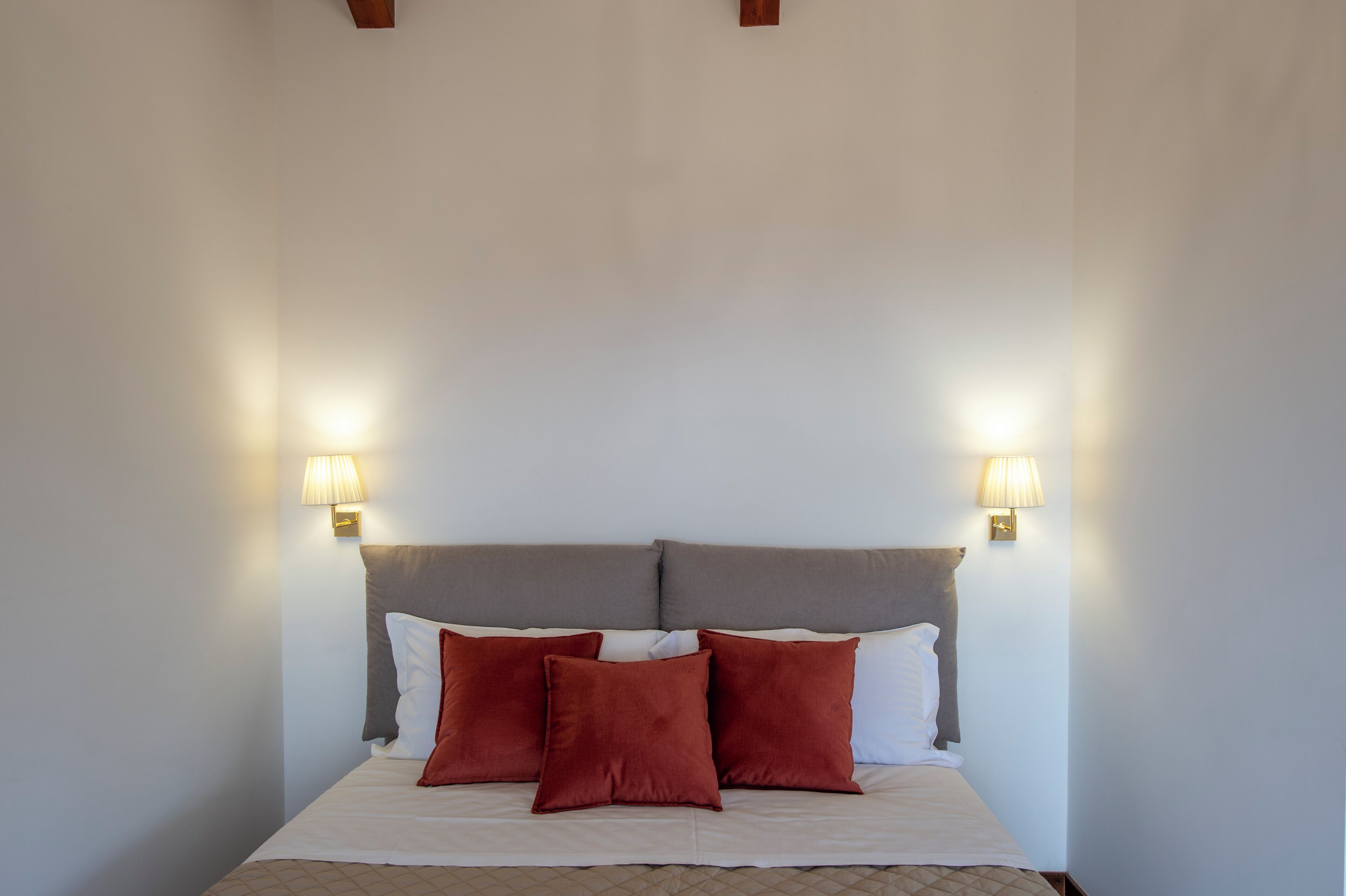 A bed with a grey headboard, white bedding, and three red pillows. There are two wall-mounted lamps on either side of the bed.