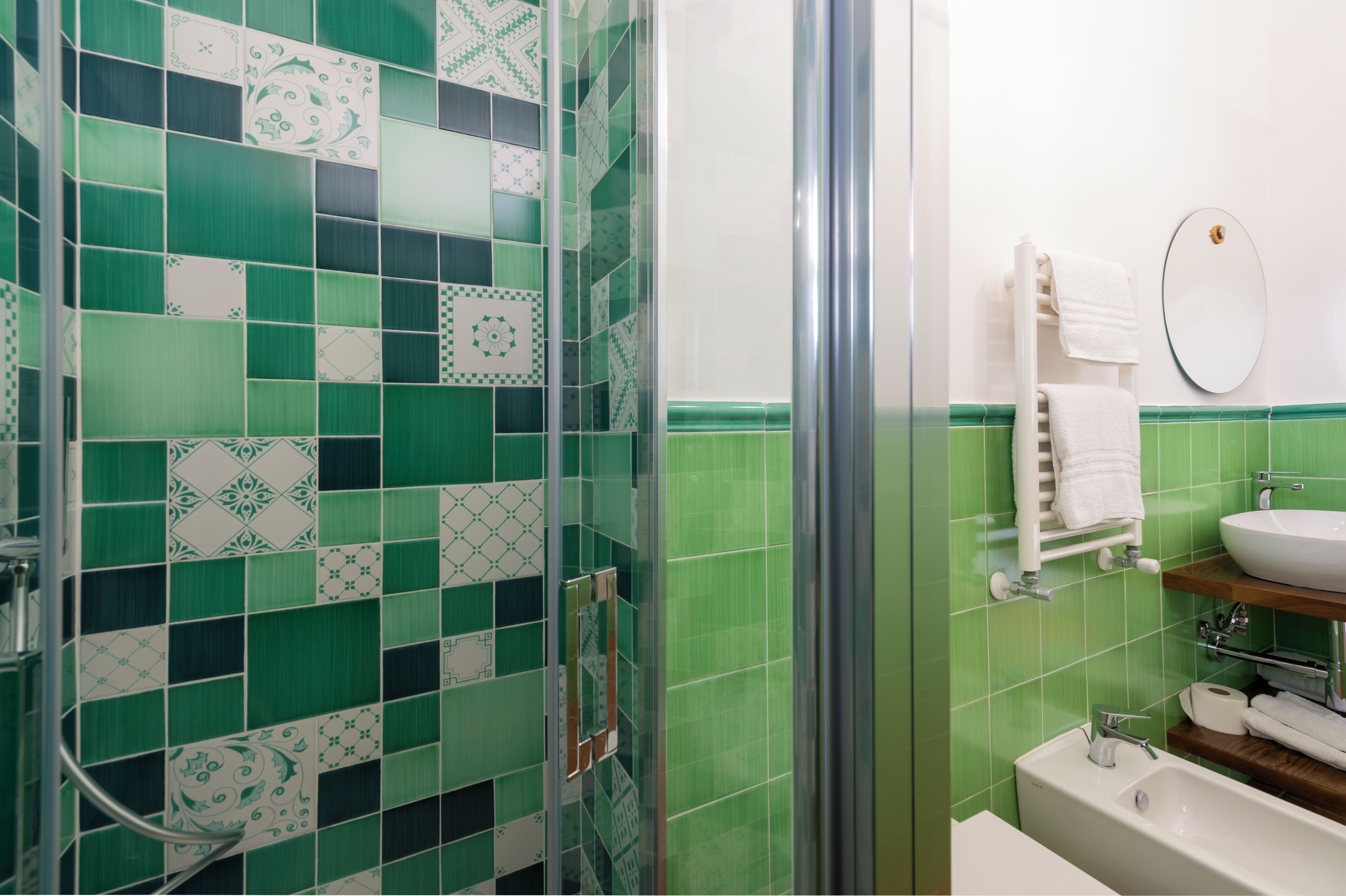 A bathroom with green and white tiles, a glass shower enclosure, a white sink, and a mirror.