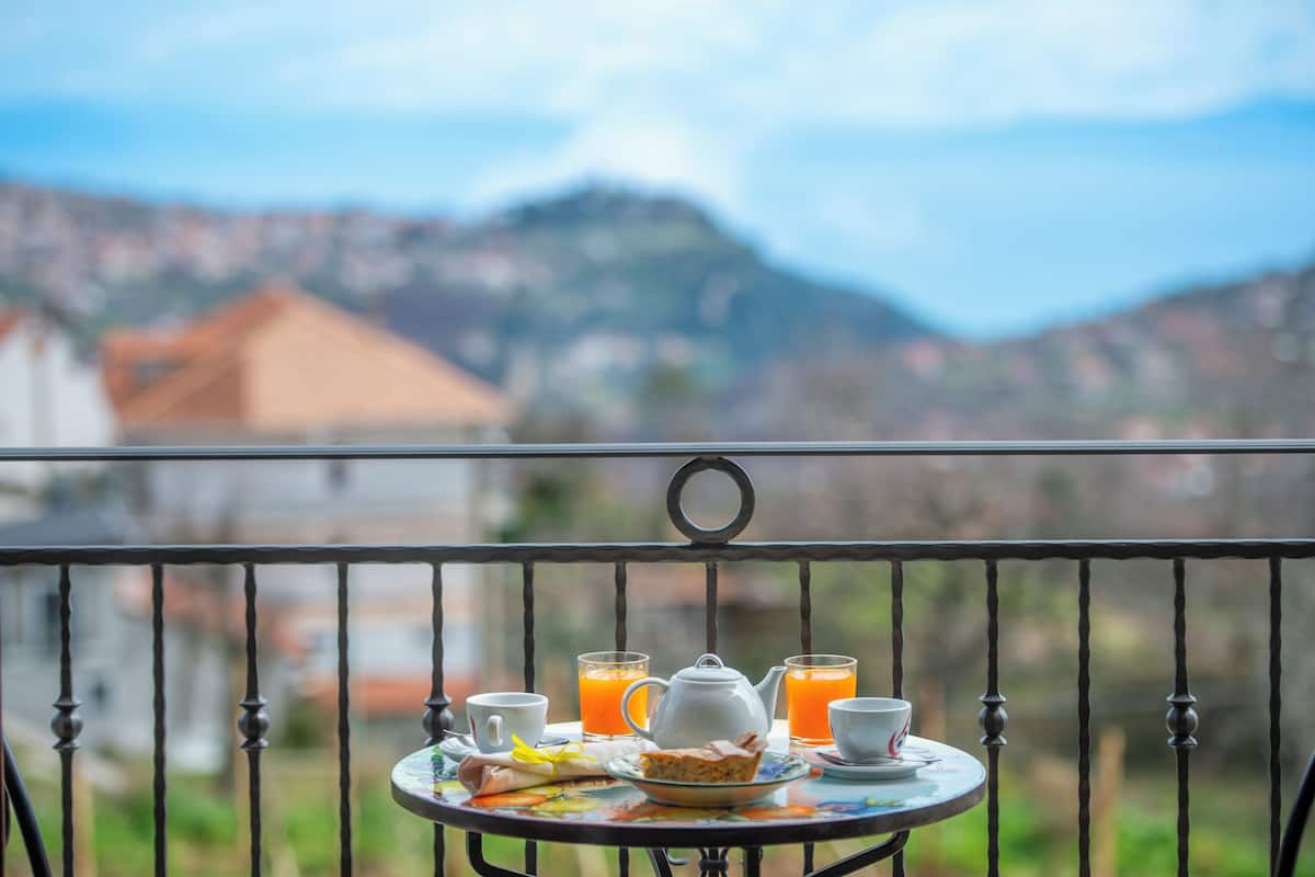 A balcony with a table set for breakfast, including a teapot, cups, and juice glasses, overlooking a mountainous landscape.