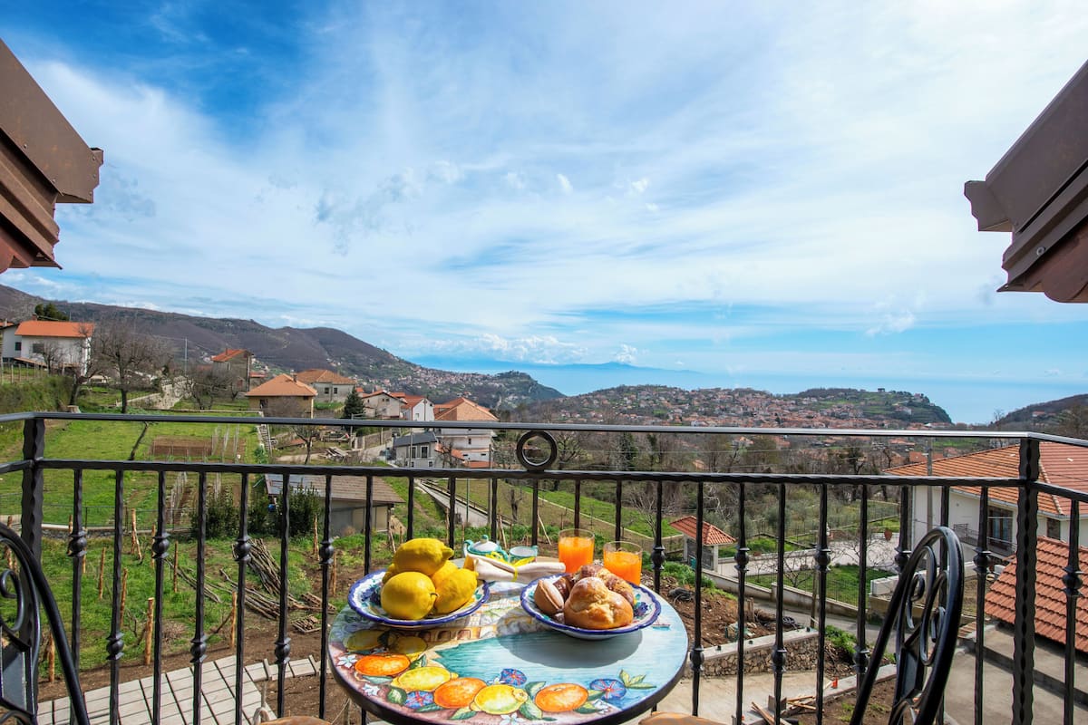 A balcony with a table set for breakfast, overlooking a scenic village and mountains.