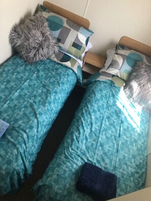 Ein kleines Zimmer mit einem Einzelbett, einem Holzkopfende, bettwäsche mit blaugrünem Muster und einem flauschigen, grauen Kopfkissen.