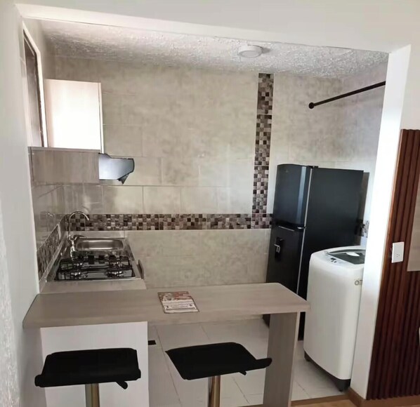 A compact kitchen with a gas stove, a sink, a refrigerator, and a small dining area with two stools.