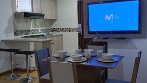 Un coin repas avec un téléviseur fixé au mur qui affiche un logo, un espace cuisine équipé d’une cuisinière à gaz et une table à manger dressée avec des assiettes et des tasses.