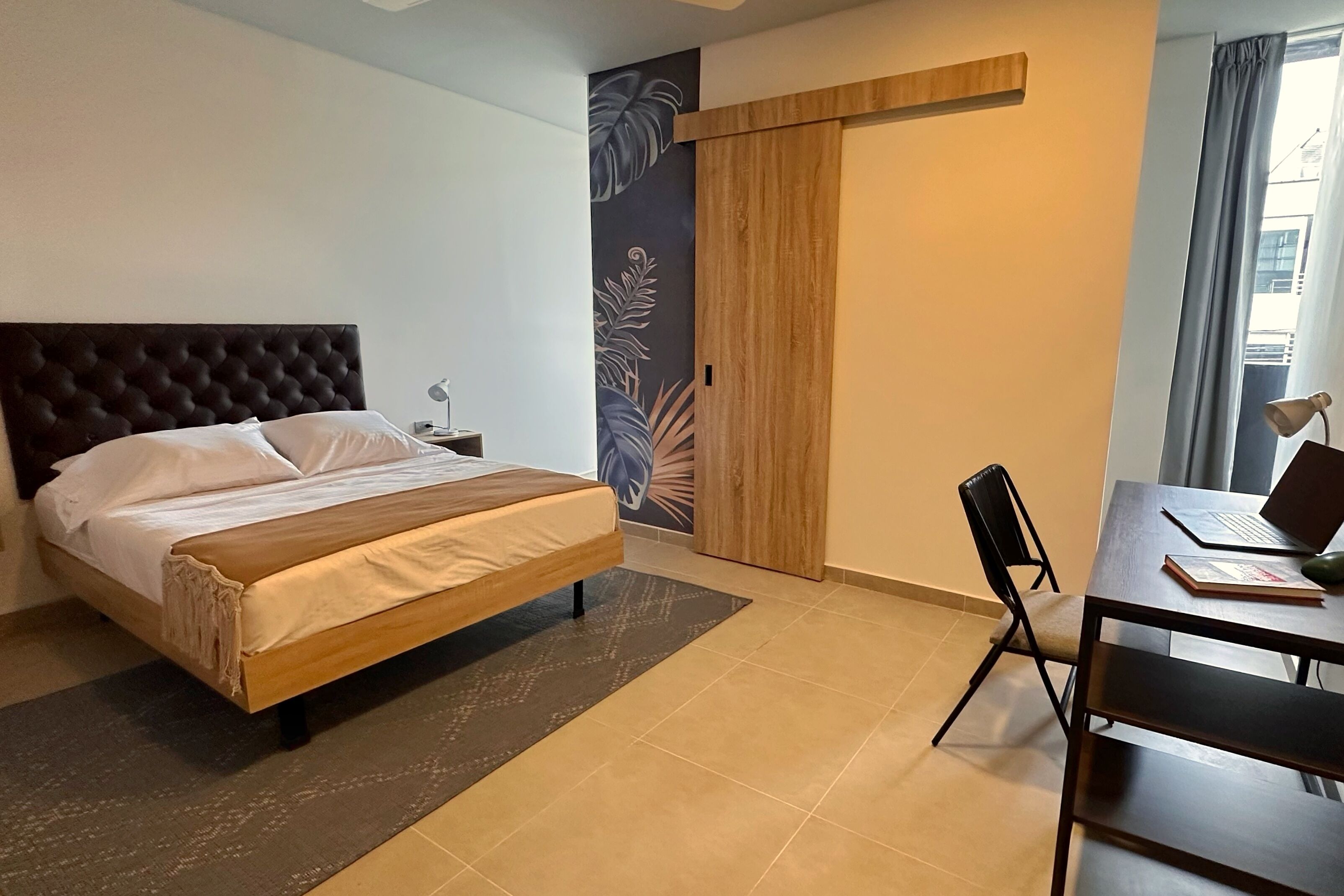 Un dormitorio con cama, un escritorio con ordenador portátil, una silla y una mesita de noche.
