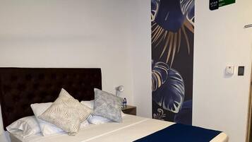 A neatly made bed with a dark blue blanket and decorative pillows, a headboard, a wall-mounted light, and a patterned rug.