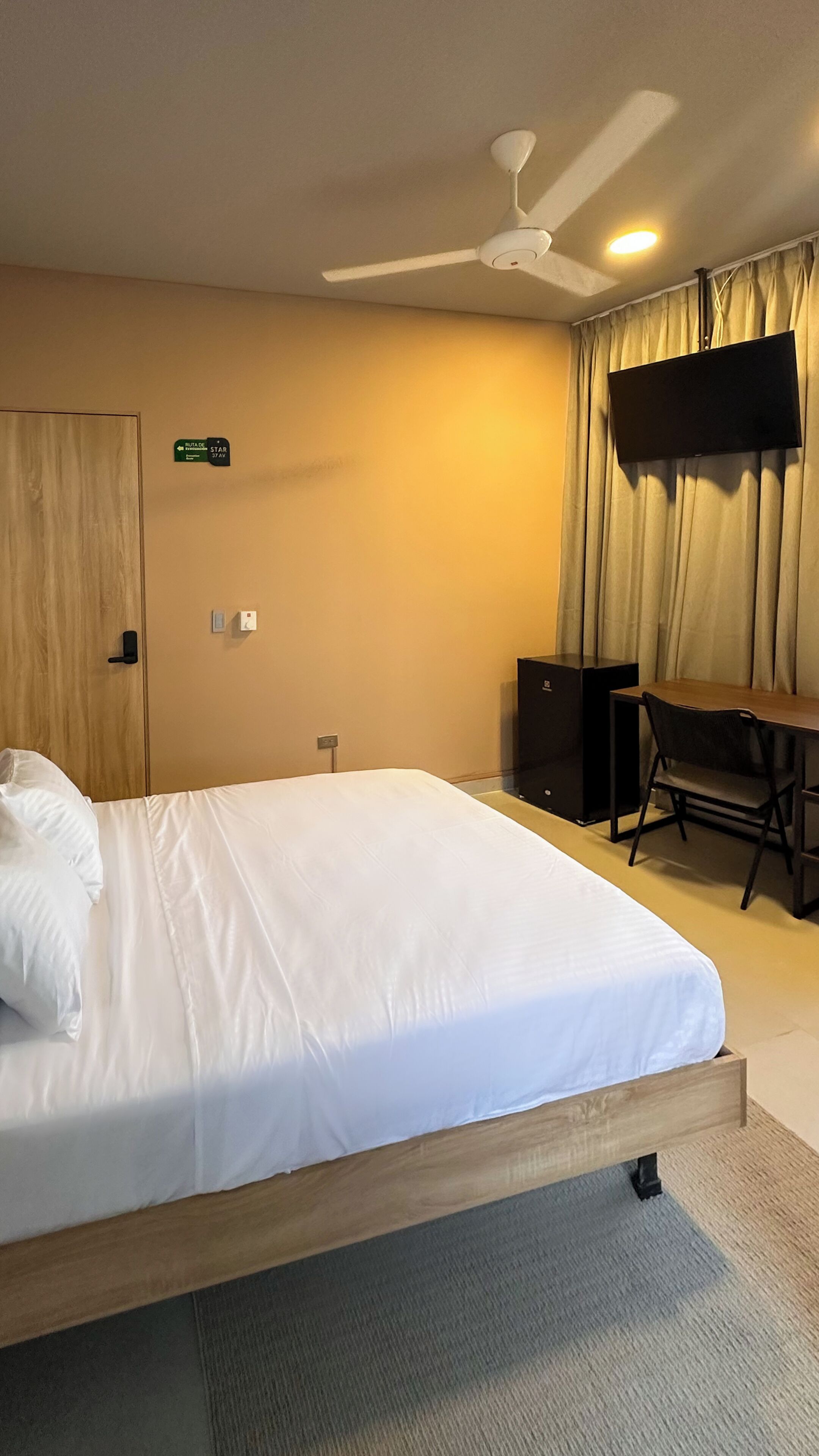 A hotel room with a bed, a desk, a chair, a television, a ceiling fan, and a sign indicating the room number.