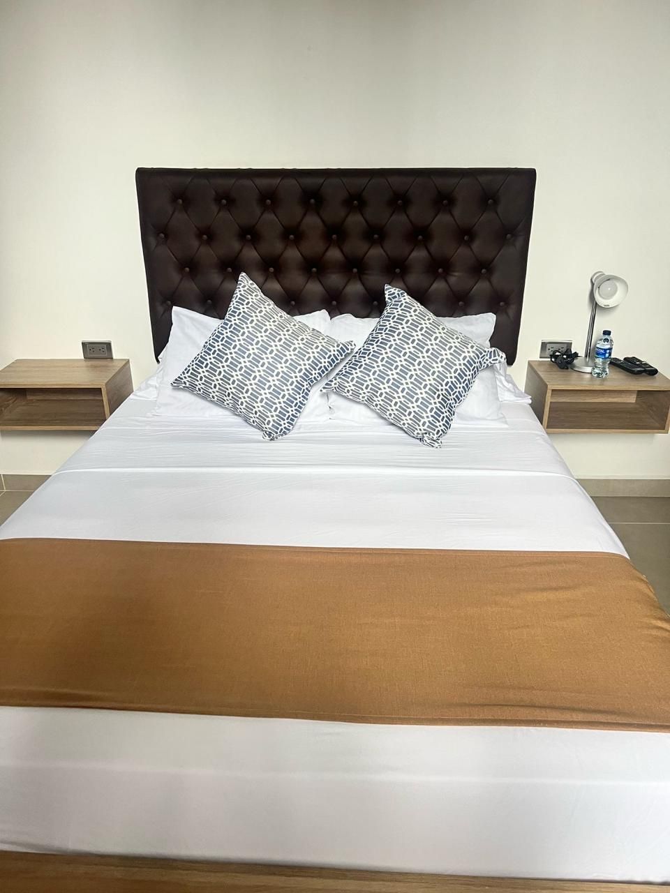 Un lit bien fait, recouvert d’une couverture marron, agrémenté de deux coussins à motifs et doté d’une tête de lit capitonnée. On y trouve deux tables de chevet : sur la première, une lampe et une bouteille d’eau, et sur la seconde, un téléphone et une télécommande.