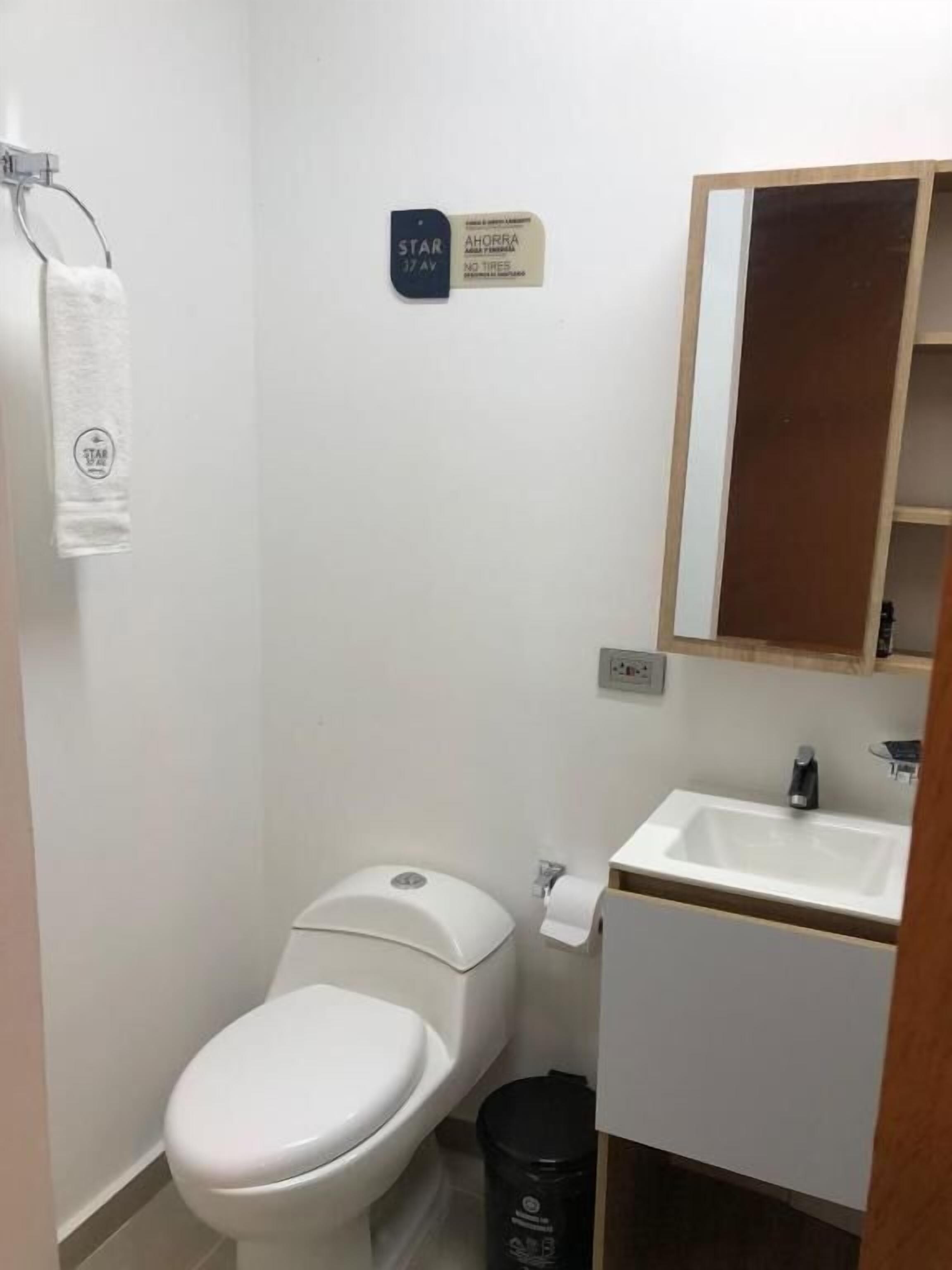 Un baño pequeño y limpio con inodoro, lavamanos y espejo. Hay una toalla colgada en un perchero y un cubo de basura.