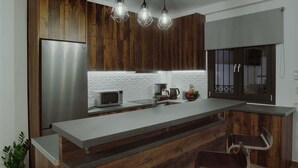 Eine moderne Küche mit Holzausstattung, einem Edelstahlkühlschrank, einer Mikrowelle und einer Kaffeemaschine. Zwei Barhocker und ein Blumentopf sind ebenfalls vorhanden.