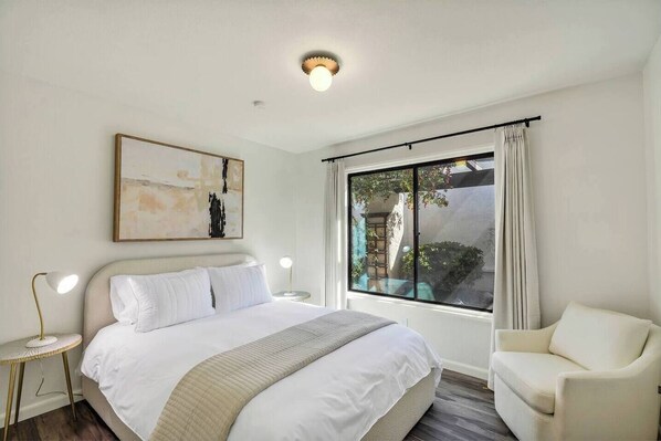 A hotel room with a large bed, a chair, a bedside table with a lamp, a window with curtains, and a ceiling light.