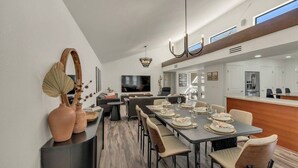 A modern dining area with a long table set for six, a kitchen in the background, and a large flat-screen TV mounted on the wall. - Desert Luxe: 3BR W/Pools, Tennis (Tucson)