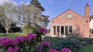 A stone house with a chimney, a porch with chairs, and a garden with pink flowers. - Canal-side 5-bed Cottage - Sleeps 10, Pet Friendly (Stoke-on-Trent)