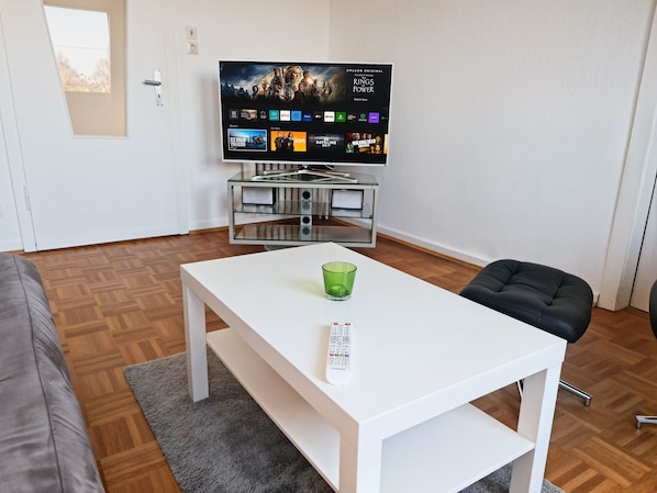 A modern living room with a flat-screen TV displaying a movie, a white table with a remote, a grey couch, and a black chair.