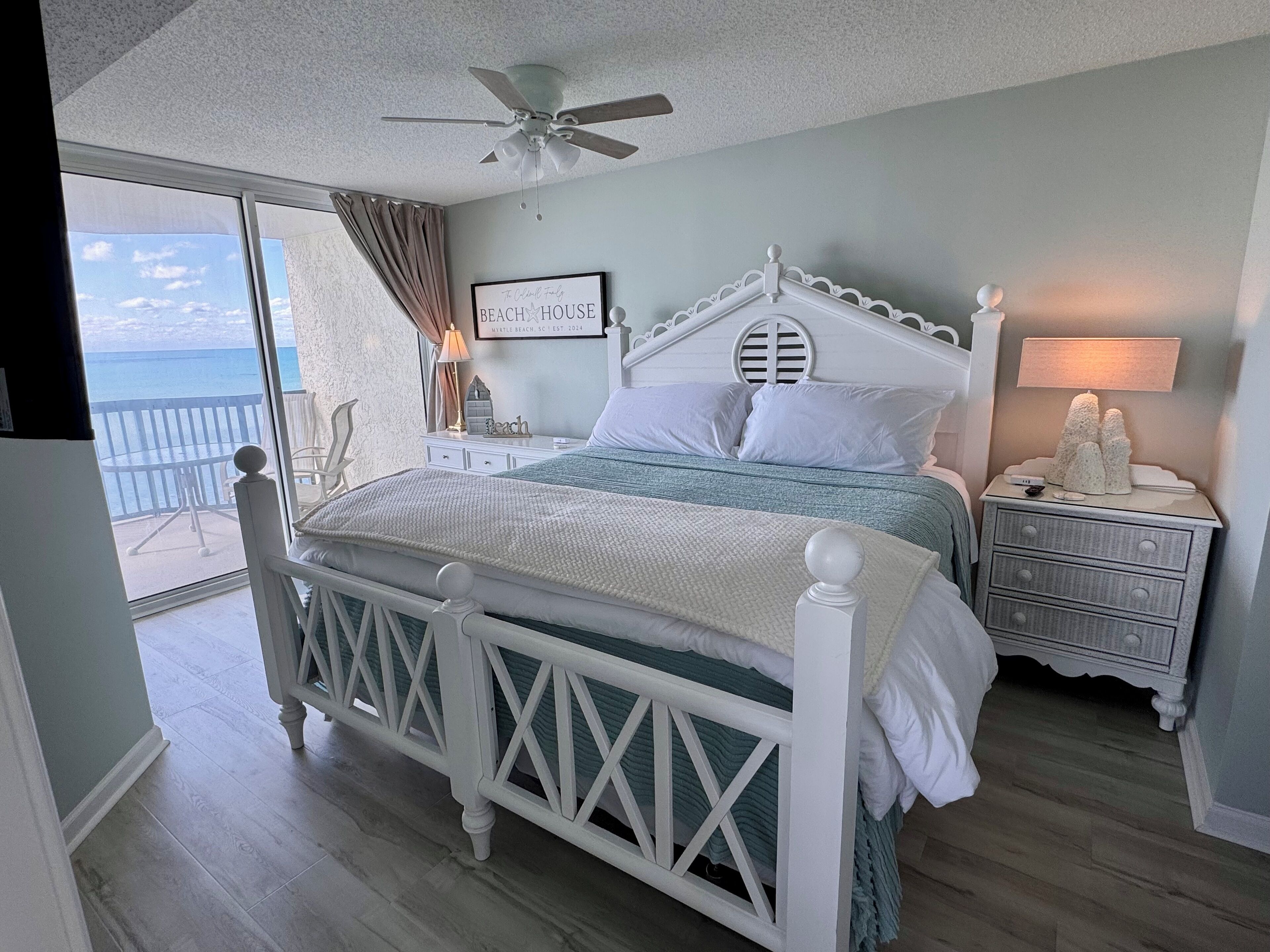 A bedroom with a white bed, a nightstand with a lamp, a ceiling fan, and a view of the ocean through a large window.