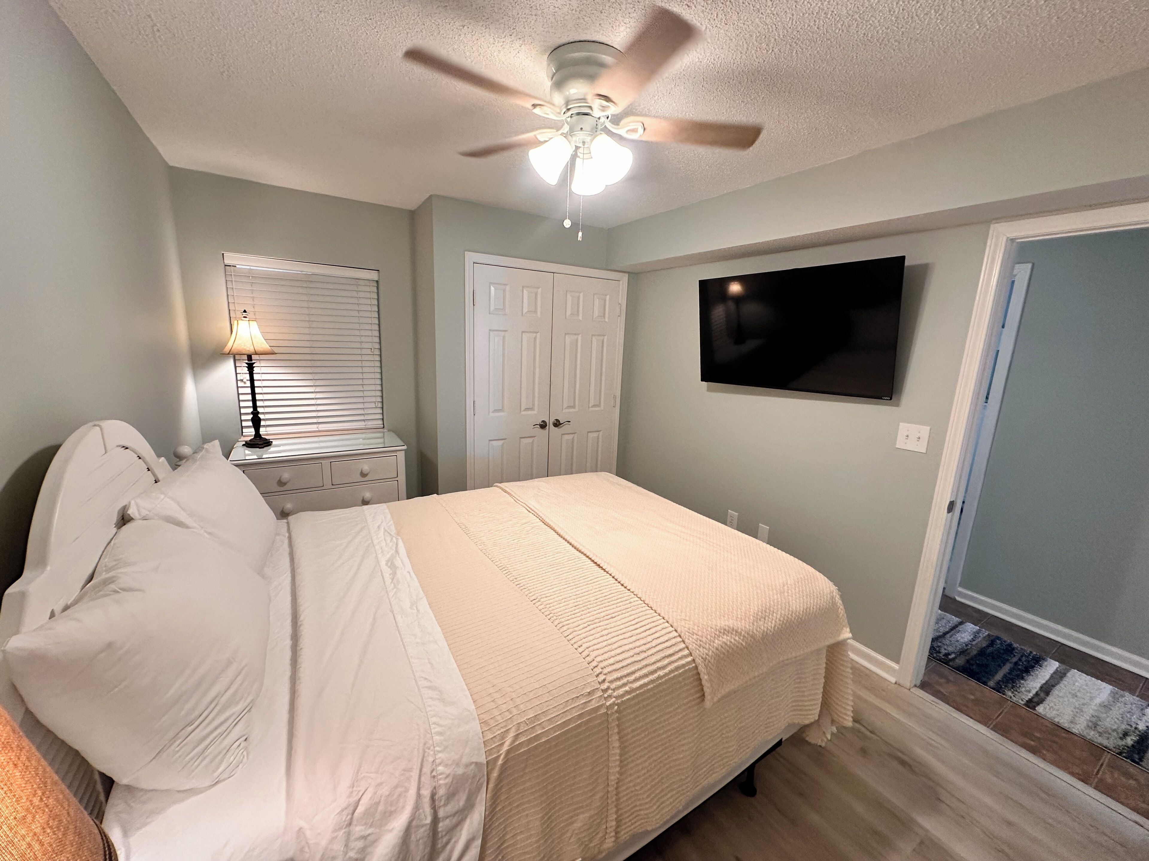 Quarto com cama, mesa de cabeceira, luminária, ventilador de teto, televisão e porta.
