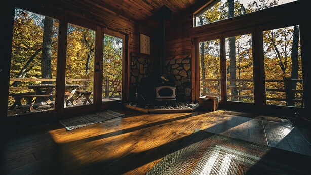 Interior de una cabaña con chimenea de piedra, piso de madera y ventanas amplias con vistas a árboles otoñales.