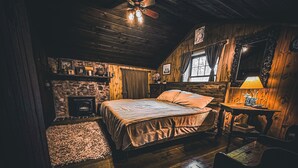 Un dormitorio de cabaña de madera acogedor, con chimenea de piedra, una cama con almohadas, una mesita de noche con lámpara y un techo de madera rústica.