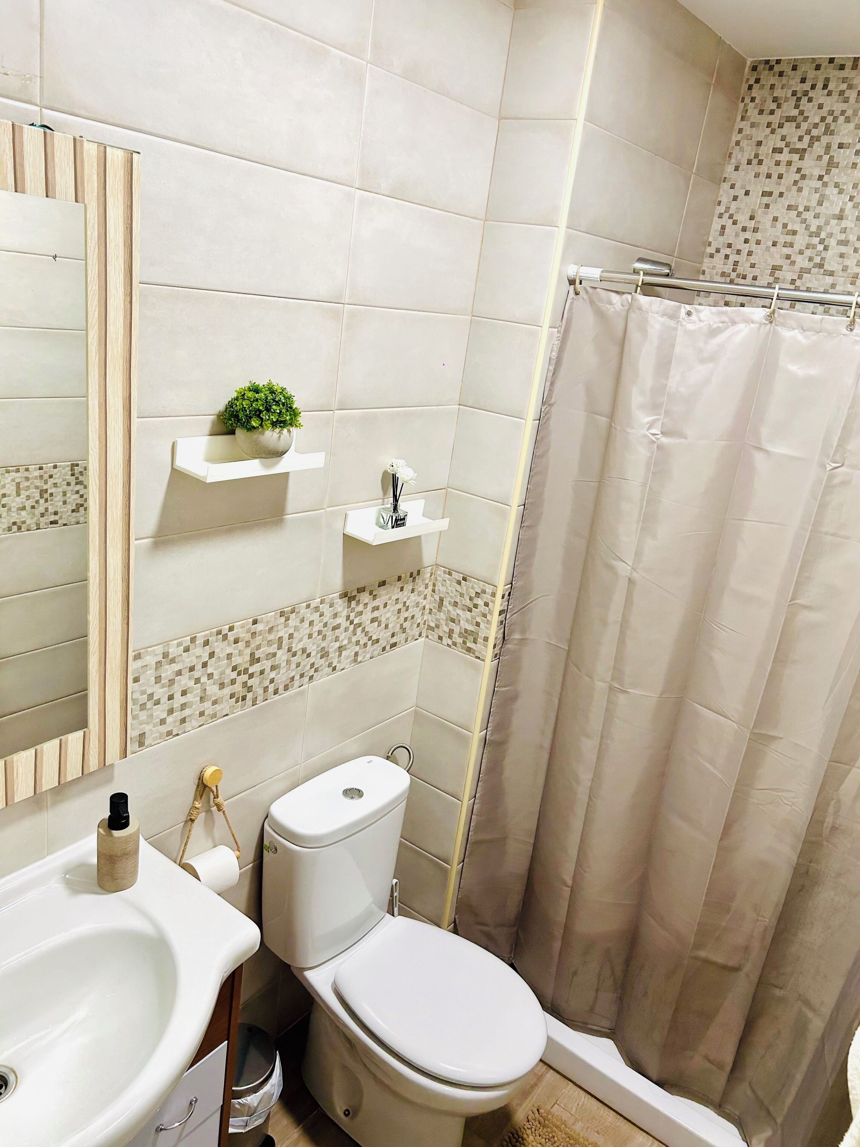 A bathroom with a toilet, sink, and shower curtain. There are two shelves with plants and toiletries.