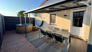 A covered outdoor area with a hot tub, a table with chairs, and a view of a building with solar panels. - Relax in the pool, whirlpool, sauna or wood-fired hot tub. (Neubörger)