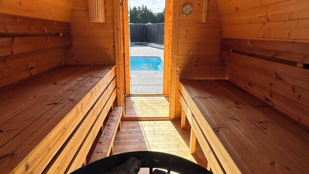 A sauna with wooden walls and benches, a bench in the foreground, and a pool visible through the doorway. - Relax in the pool, whirlpool, sauna or wood-fired hot tub. (Neubörger)