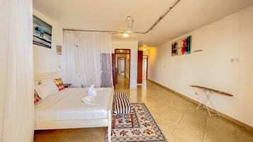 A hotel room with a bed, a bedside table, a chair, a small table, a curtain, a painting, and a hallway leading to another room.