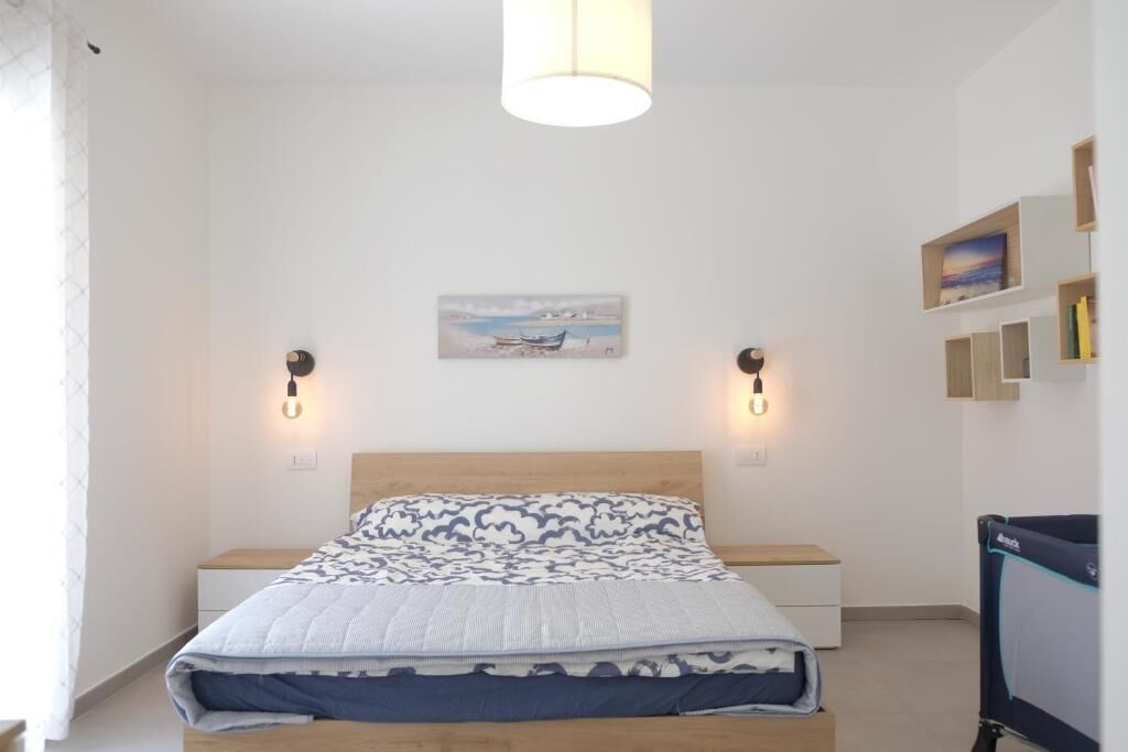 A neatly arranged bedroom with a bed, bedside tables, a wall-mounted lamp, and a small shelf with books.