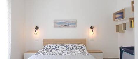 A neatly arranged bedroom with a bed, bedside tables, a wall-mounted lamp, and a small shelf with books.