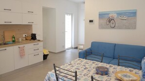 Et moderne køkken med hvide skabe og en træbordplade, et spiseområde med en blå sofa og et bord med dug med et mønster, samt et maleri med et strandmotiv på væggen.