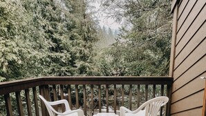 A balcony with two white plastic chairs and a table, surrounded by dense evergreen trees.