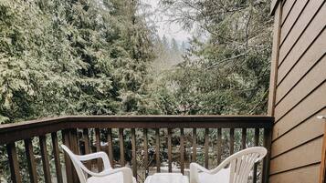 A balcony with two white plastic chairs and a table, surrounded by dense evergreen trees.