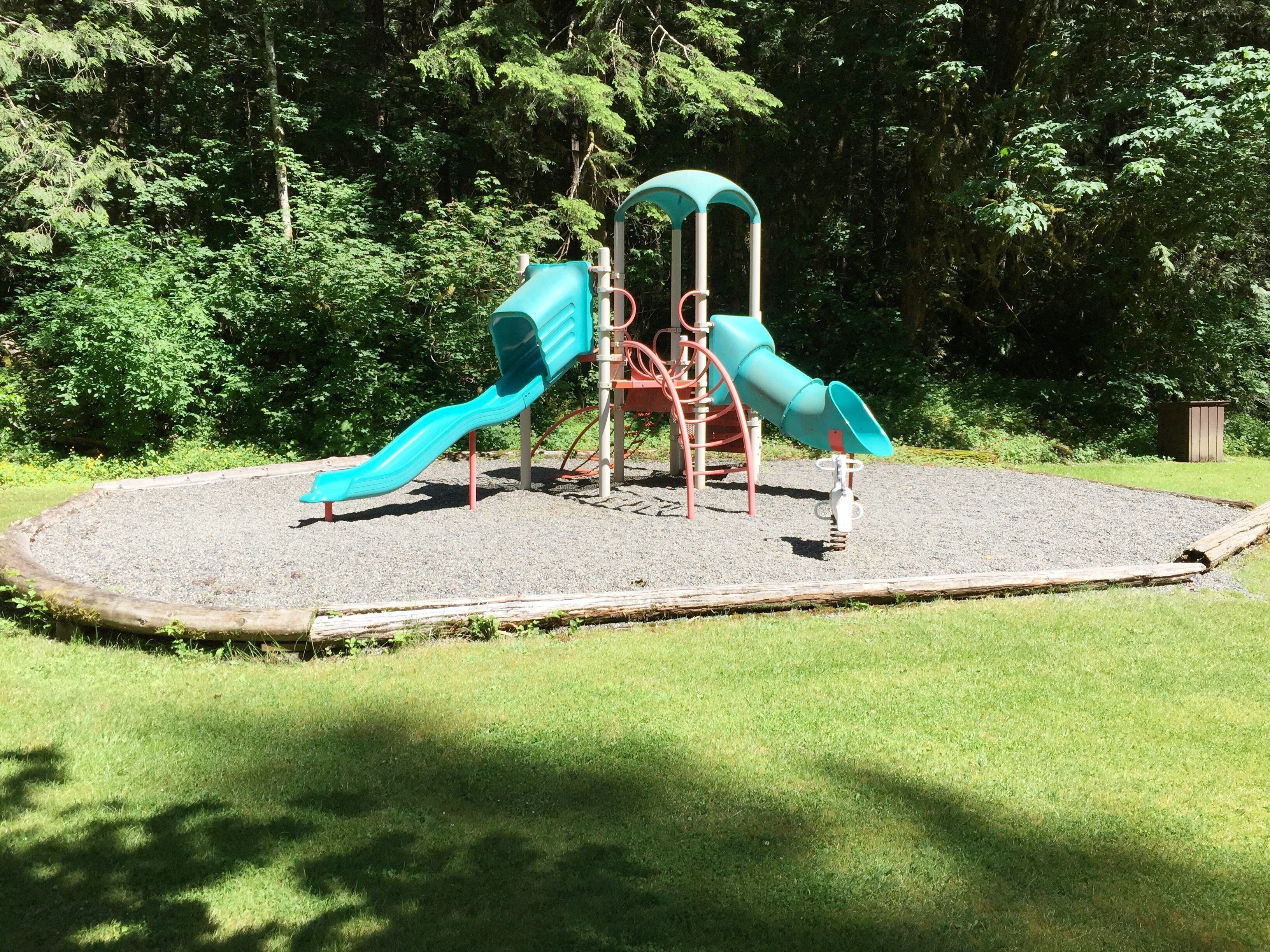 A playground with two slides and a climbing structure, surrounded by a grassy area and trees.
