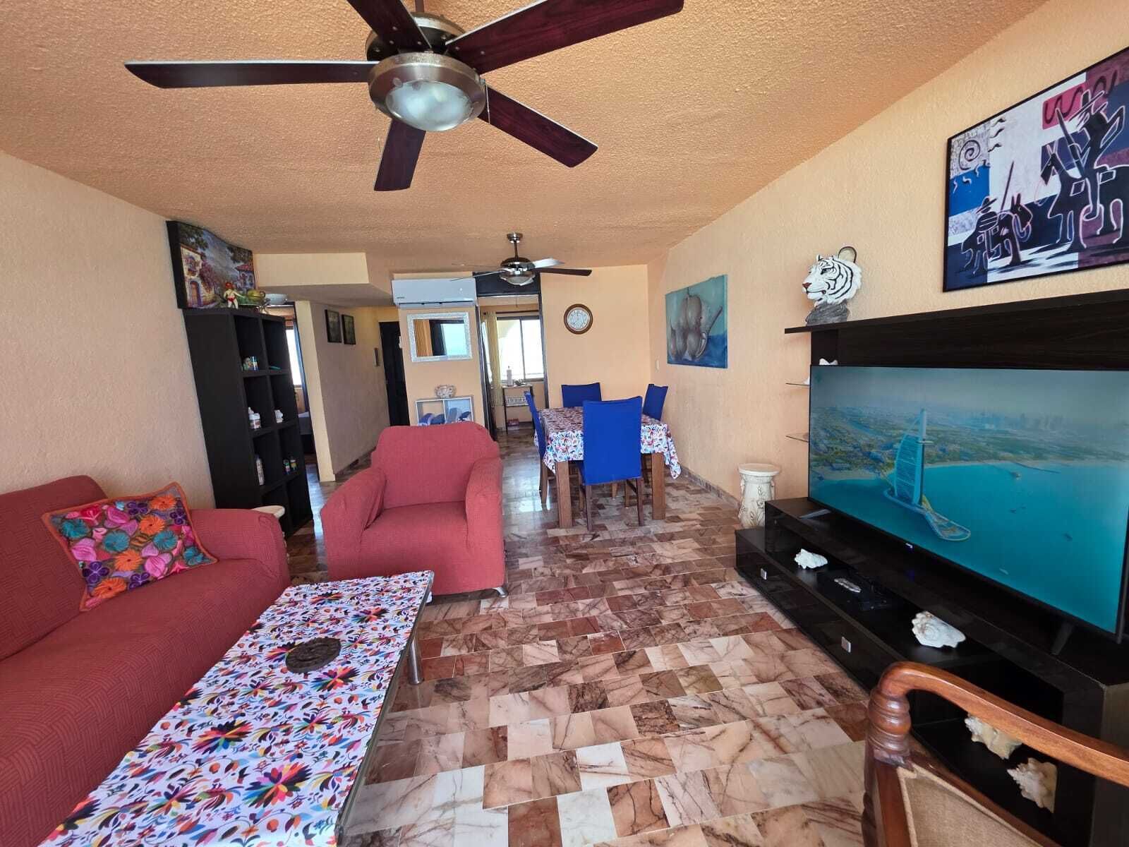Un salon avec un téléviseur, un canapé rouge, une table à motifs, un ventilateur de plafond et un coin repas avec des chaises bleues.