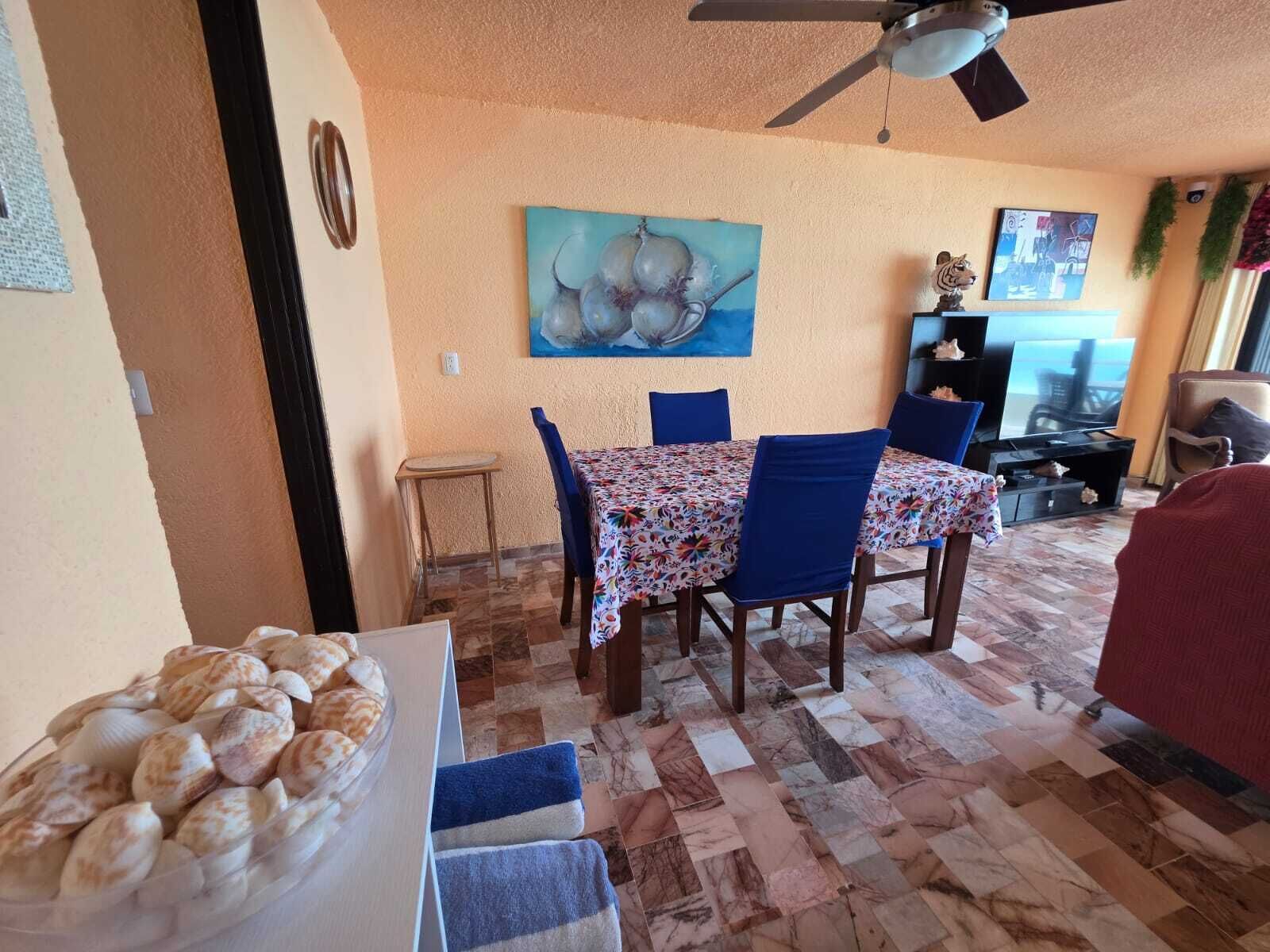 Un coin repas avec une table dressée pour quatre personnes, un ventilateur de plafond et un tableau accroché au mur.