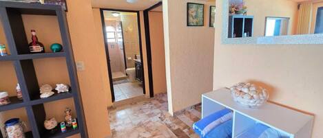 A hotel room with a tiled floor, a shelf with decorative items, a bathroom visible through an open door, and a wall-mounted air conditioner.