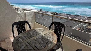A balcony with a wooden table and two chairs, overlooking a beach with turquoise waters and a cityscape in the distance. - Ocean view condo with balcony hotel zone (Cancún)