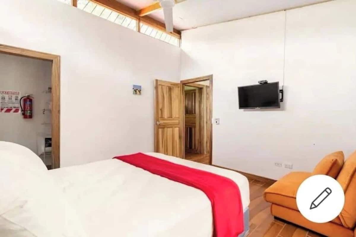 Ein Hotelzimmer mit einem Bett, einer roten Decke, einem Fernseher, einem orangefarbenen Sessel und einer Holztür.