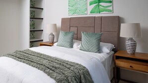 Una cama bien hecha con una manta verde, dos almohadas y un cabecero. Hay dos mesitas de noche con lámparas y un tocador con una lámpara. Se observa un armario a la izquierda.