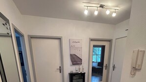 Een gang met houten vloer, witte wanden en een plafondlamp. Er hangt een lijst met een afbeelding aan de muur en er staat een klein zwart console tafeltje.