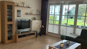 Een woonkamer met een televisie, een houten kast en een raam met uitzicht op groen.