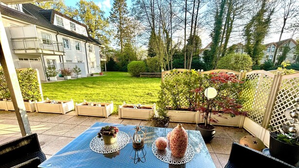 Een terras buiten met een tafel voor twee personen, omringd door potplanten en een uitzicht op een verzorgd gazon en een huis op de achtergrond.