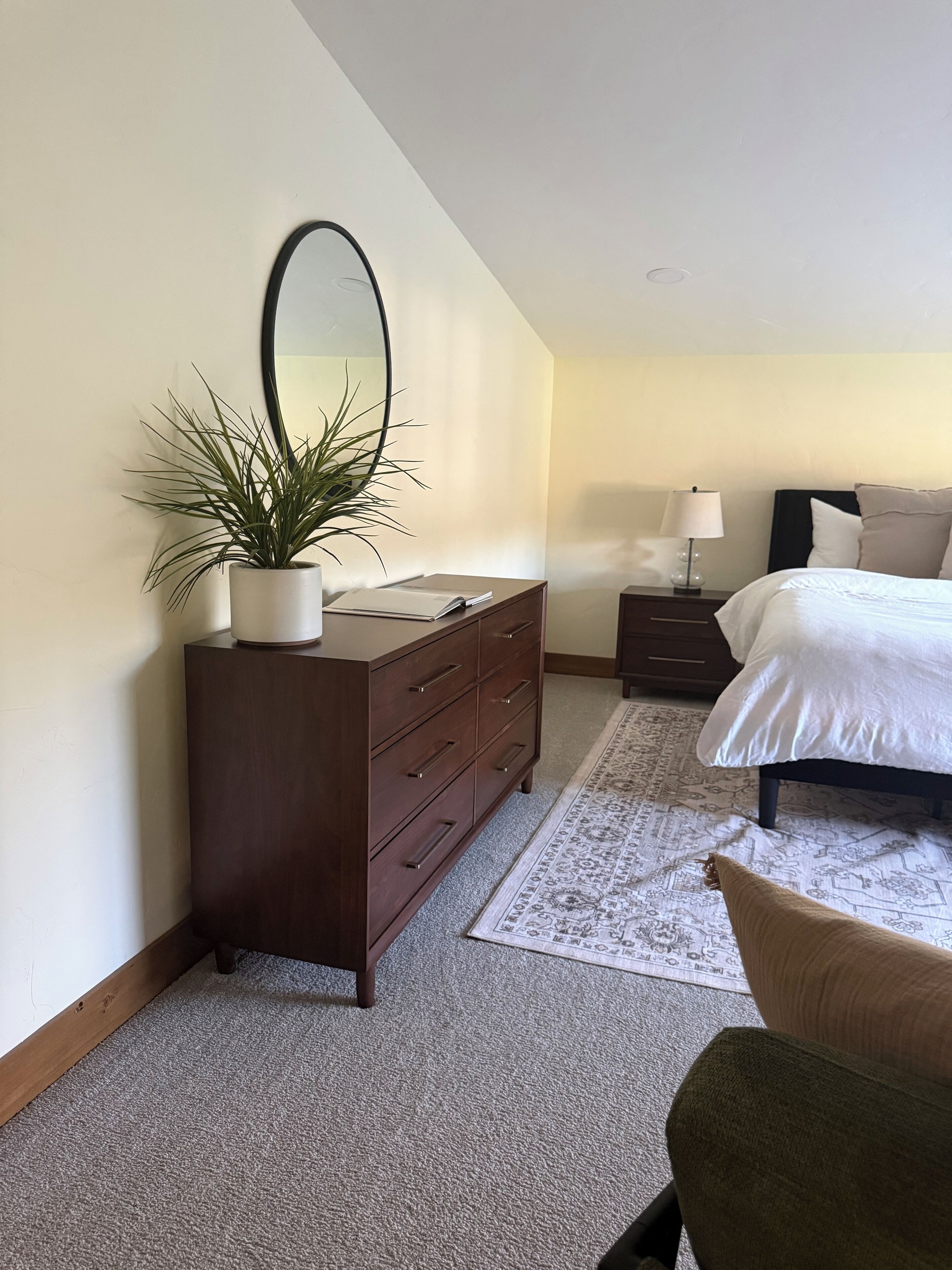 A bedroom with a large bed, a dresser with a mirror, a bedside table with a lamp, and a potted plant.