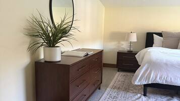 A bedroom with a large bed, a dresser with a mirror, a bedside table with a lamp, and a potted plant.