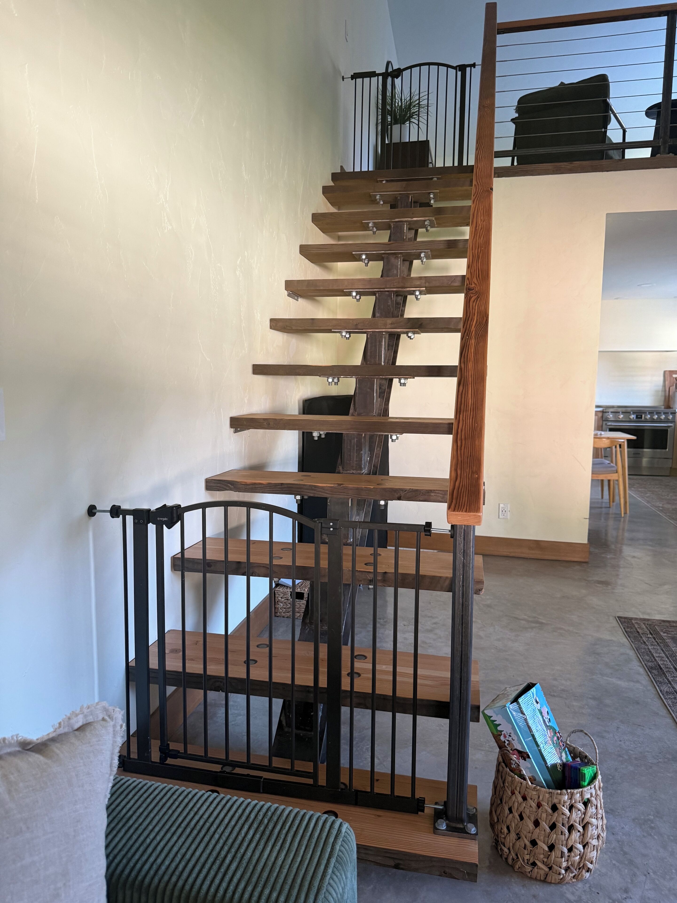 Una scala a chiocciola in legno con ringhiere in metallo, un cesto di vimini sul pavimento e una zona cucina sullo sfondo.