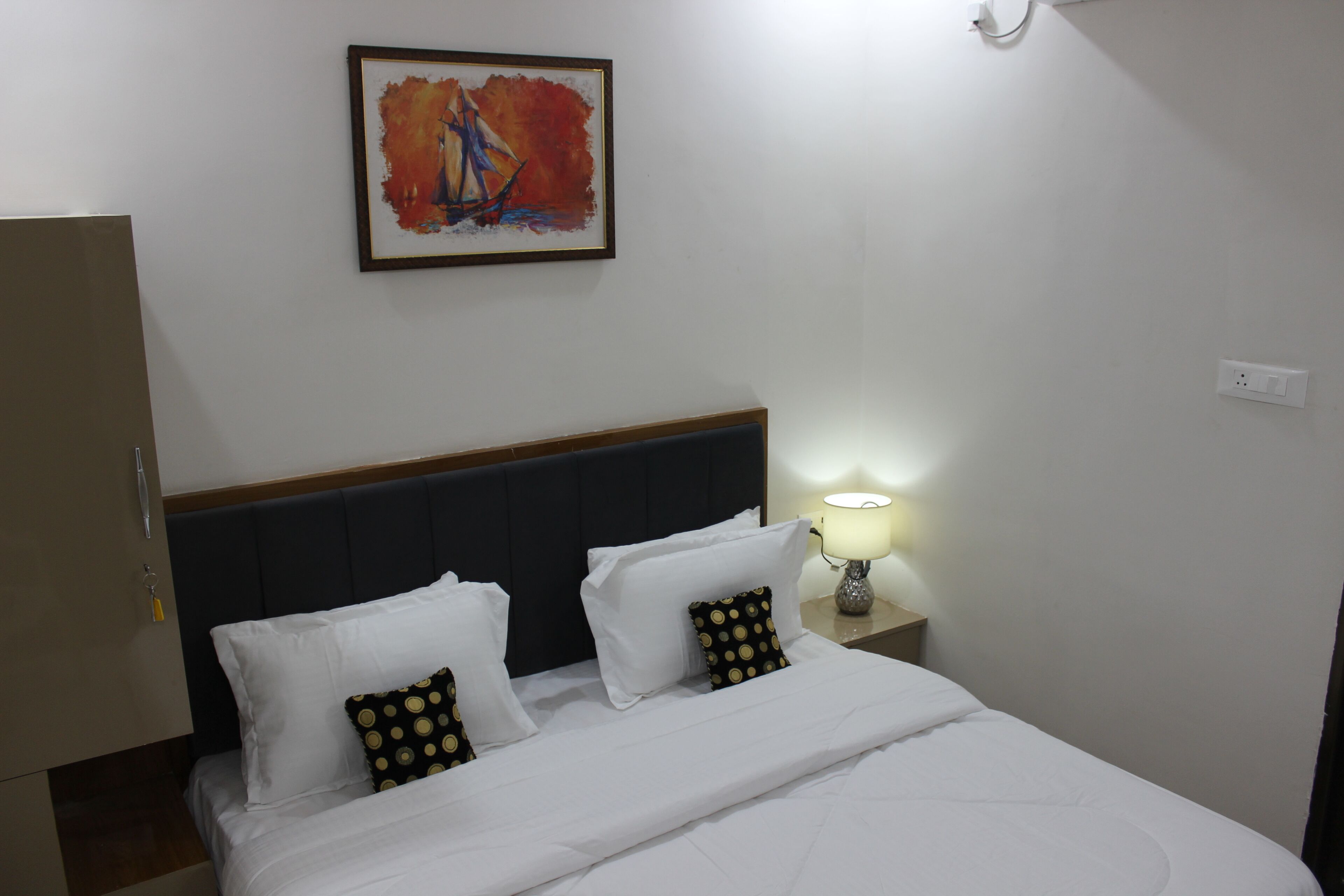 A neatly made bed with white linens and two decorative pillows, a bedside table with a lamp, and a framed painting above the bed.