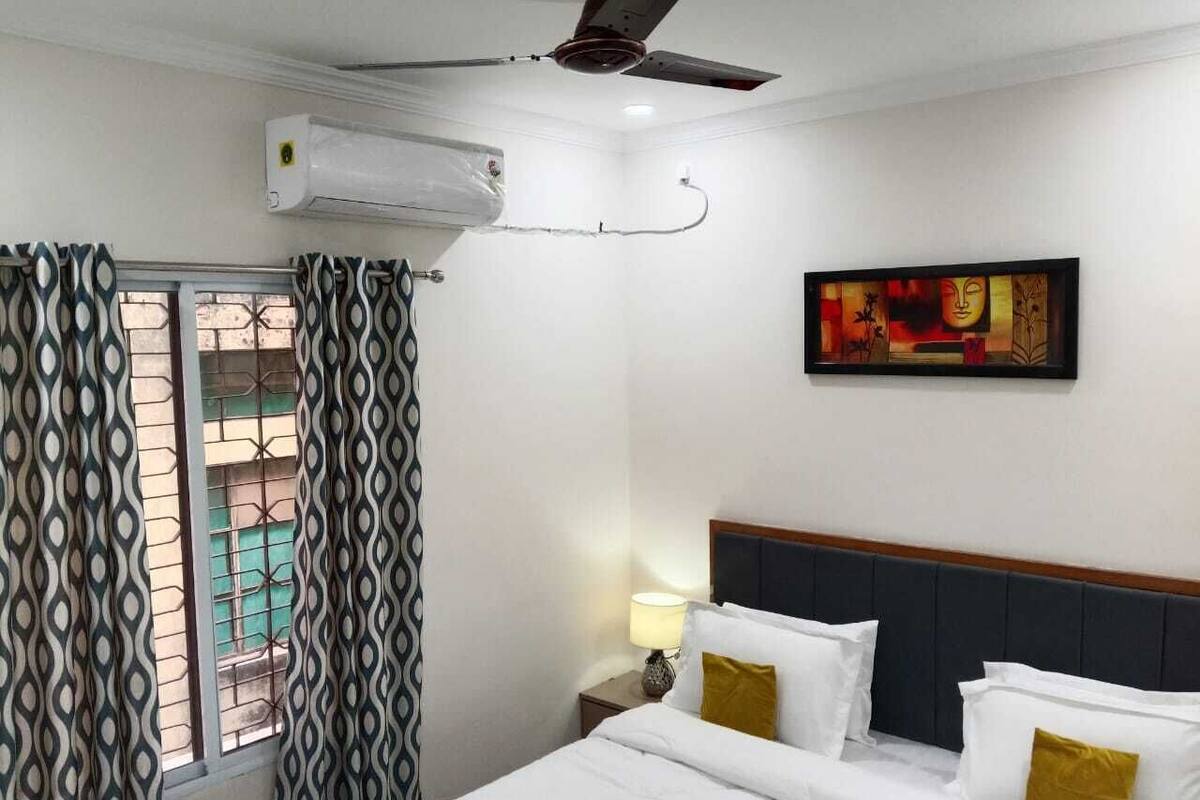 Egy szállodai szoba, amelyben található egy ágy, egy mennyezeti ventilátor, egy légkondicionáló, egy mintás függönnyel ellátott ablak, egy éjjeliszemélyzet, rajta egy lámpával, és egy falra szerelt festmény.