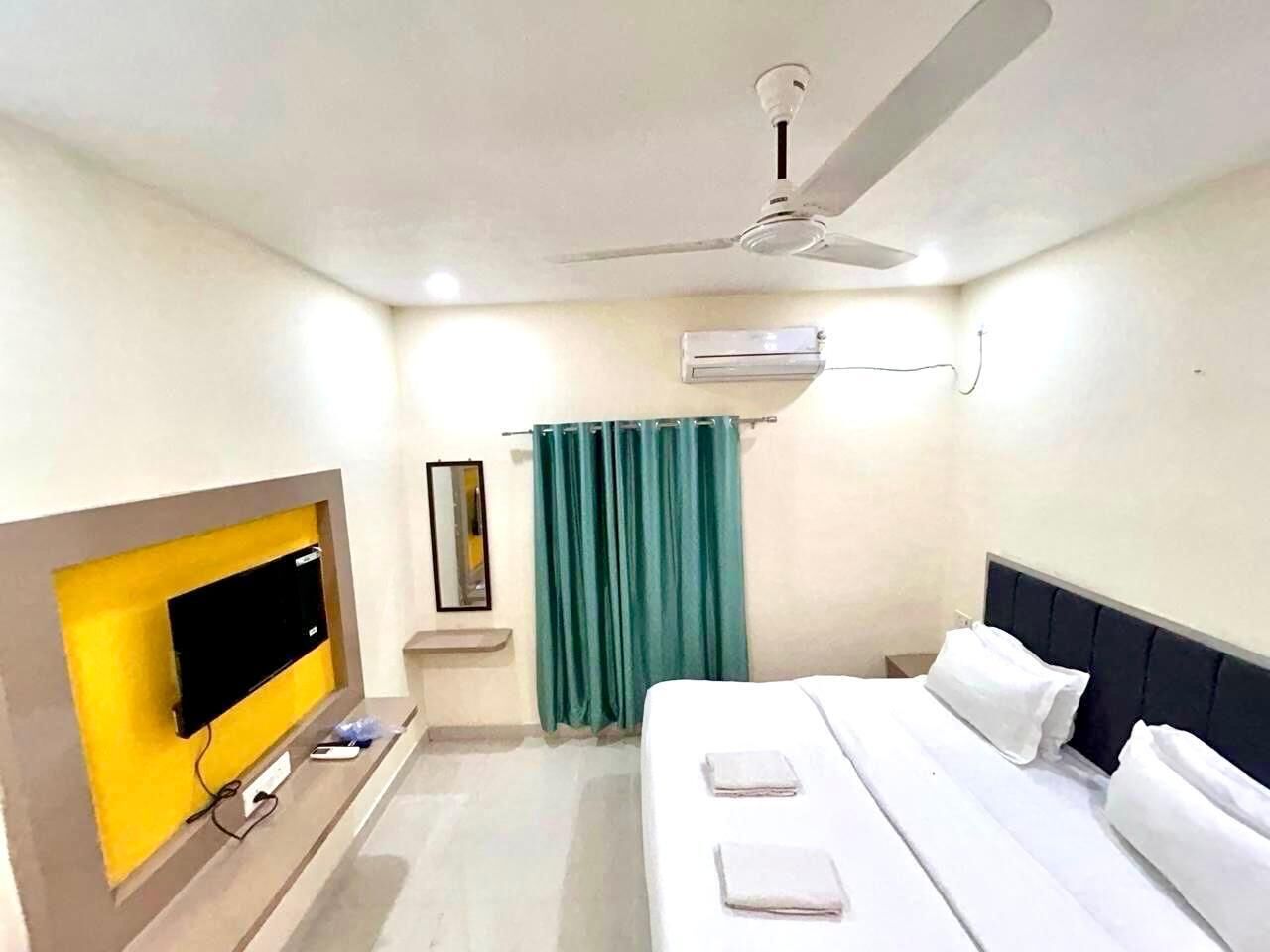 A hotel room with a bed, a TV mounted on a yellow wall, a ceiling fan, and a window with green curtains.