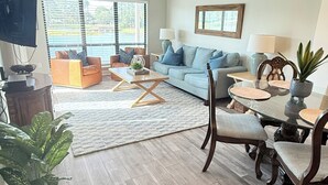 Una sala de estar con sofá, mesa de centro y mesa de comedor con sillas. Hay un televisor montado en la pared y una ventana con persianas.