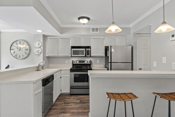 A modern kitchen with white cabinetry, stainless steel appliances, a built-in oven, and a microwave. There are two pendant lights, a clock, and a small island with bar stools. - Charming 2-bedroom condo in desirable Sandestin Resort, GOLF CART INCLUDED!! (Destin)