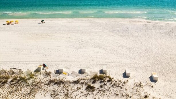Una playa con arena blanca y agua azul cristalina. Hay sombrillas y sillas de playa a lo largo de la orilla.