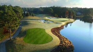 Un campo de golf con una masa de agua a un lado, una carretera que serpentea alrededor del campo, y un edificio al fondo.